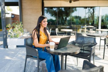Bir kadın gülümsüyor, dışarıda bir kafede dizüstü bilgisayarda yazıyor, gün boyunca uzaktan çalışmaktan ve kahveden zevk alıyor.