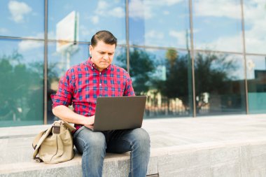 Bilgisayarda yazı yazan, esnek bir program ve açık hava çalışma alanının keyfini çıkarırken işine ya da derslerine odaklanan sıradan giyinmiş bir adam.