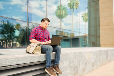 İspanyol adam dizüstü bilgisayarına odaklanmış, çağdaş bir binanın dışında otururken daktilo yazıyor, kentsel yaşam tarzını ve teknolojiyi yansıtıyor.