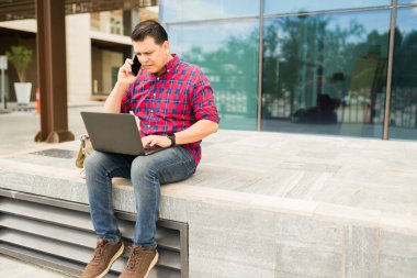 Yetişkin bir adam stresli ve endişeli hissediyor, dışarıda dizüstü bilgisayarda yazarken cep telefonuyla konuşuyor.
