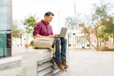Parkta dizüstü bilgisayar kullanan, gülümseyen ve konsantre olan bir adam. Uzaktan çalışan, dijital göçebe ya da dışarıda okuyan öğrenci