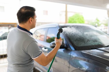 Arabasını güçlü bir hortum hortumuyla iyice yıkayan ve garajda araç bakımını yapan bir adamın arka görüntüsü.