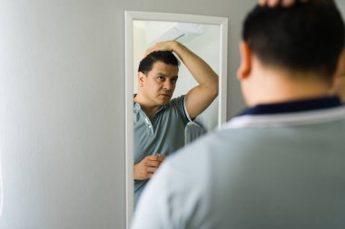 Bir adam saçını şekillendirirken yansımasına bakıyor, evde kişisel bakımına ve fiziksel görünüşüne odaklanıyor.