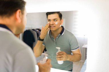 Banyodaki aynanın önünde duran mutlu adam günlük cilt bakımının bir parçası olarak yüzüne nemlendirici krem sürüyor.