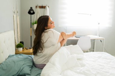 Şafak vakti yatakta uzanan iri yarı bir kadın, çarşafların üzerinde güneş ışığı, rahat ve rahat bir pijamayla, yavaş ve sağlıklı bir sabah rutinini kucaklayan bir kadın.