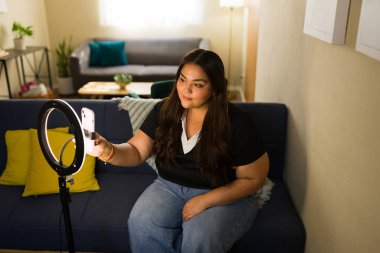 Genç içerik yaratıcısı sosyal medya için video kaydediyor evdeki koltukta oturuyor, cep telefonuyla canlı yayın yapıyor.