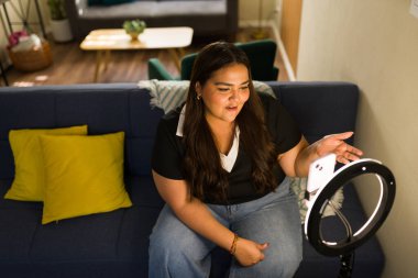 Genç bayan içerik yaratıcısı akıllı telefondan canlı yayında ev koltuğundan, halka ışığı altında hareketli ve çekici takipçiler