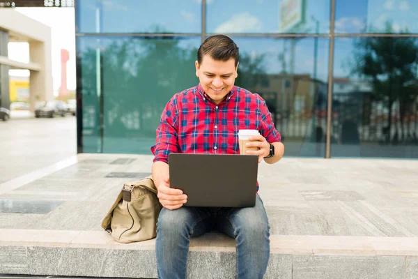 Genç adam dışarıda oturuyor, dizüstü bilgisayarı üzerinde çalışıyor ve kahve içiyor, uzaktan çalışmayı, internetten öğrenmeyi ve dijital göçebe yaşam tarzını temsil ediyor.