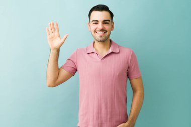 Gülümseyen ve el sallayan genç bir adam mavi arka planda dostça bir jest yapıyor.