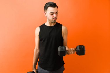 Adam dambılla biseptik kıvrım egzersizi yapıyor, turuncu bir arka plan üzerinde vücut çalışmasına konsantre oluyor.