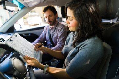 Kadın öğrenci, ehliyeti için bir kontrol listesi hazırlıyor. Erkek sürücü eğitmeniyle birlikte bir araçta.