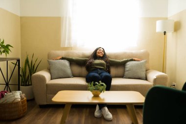 Latin bir kadın evinin oturma odasında bir kanepede oturuyor, kollarını uzatıyor ve gözleri kapalı gülümsüyor, rahatlık ve rahatlığın tadını çıkarıyor.