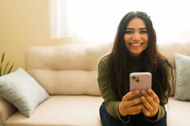 Neşeli İspanyol kadın cep telefonu tutuyor ve evinde rahat bir kanepede otururken gülümsüyor.