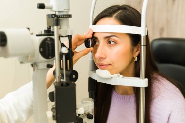 Modern bir klinikte rutin görme sağlığı için kesik lamba kullanan bir göz doktorunun göz muayenesi yaptıran Latin bir kadın.