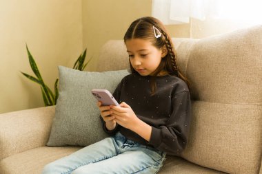 Oturma odasındaki kanepede rahatlayan genç bir kız, cep telefonu kullanıyor. Cihazındaki dijital içerikle meşgul.