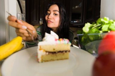 Latin bir kadın buzdolabının içine uzanıyor, çatal dolusu lezzetli pasta alıyor ve koyu renkte serpiştiriyor.