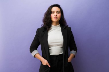 Profesyonel genç iş kadını bir stüdyoda duruyor, siyah ceket ve beyaz bir bluz giyiyor, özgüven ve profesyonelliği canlandırıyor.