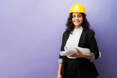 Gülümseyen kadın mühendis proje planları yapıyor, başlık takıyor, teknik bir kariyerde başarı ve profesyonelliği temsil ediyor.