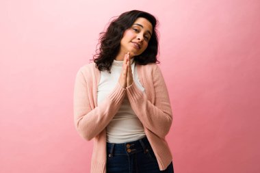 Tatlı bir kadın suçlu ve pişman görünüyor, dua etmek için el ele tutuşuyor, pembe arka planda yardım istiyor.