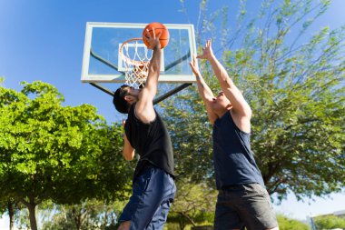 Parkta basketbol oynayan iki genç adam, bir oyuncu topa vururken diğeri onu engellemeye çalışıyor.
