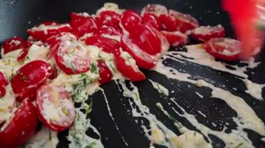 Erimiş peynir ve aromatik otlarla karıştırılmış taze vişneli domatesler siyah bir tavada görsel olarak çekici bir mutfak sahnesi yaratır. Canlı kırmızı ve kremalı dokular yemek pişirmek için harika bir ilham kaynağı sunuyor..