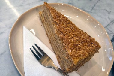 Çekici bir dilim bal pastası seramik bir tabakta, katmanlı sünger ve ekmek kırıntılarını sergiliyor. Bir çatal ve peçeteyle birlikte bu tatlı, hoşgörülü bir ikram ya da tatlının tatlısı için mükemmeldir..