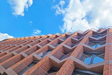 Canlı, mavi bulutlu bir gökyüzüne karşı çağdaş bir tuğla binanın görsel açıdan çarpıcı alçak açılı görüntüsü. Bu mimari görüntü geometrik desenleri ve modern tasarımı vurgular, şehir ve şehir temaları için mükemmeldir.