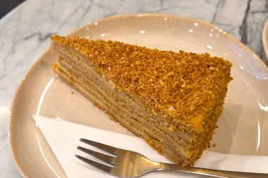 Bir dilim bal katmanlı kek ve üzeri çıtır çıtır ekmek mermer bir masada beyaz bir tabakta duruyor. Yanında kağıt peçetenin üzerinde bir çatal, tatlı meraklıları için mükemmel bir tatlı sergiliyor..