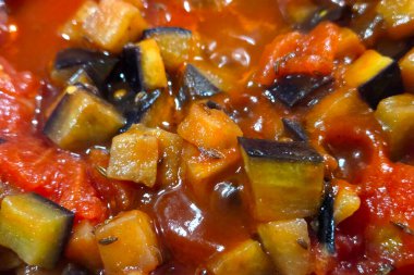 Canlı, baharatlı bir sosun içine yerleştirilmiş patlıcan ve domates küplerinin yer aldığı zengin renkli bir ratatouille çanağının yakın plan görüntüsü. Dokular ve renkler bu klasik provenkal tarifin rustik cazibesini ve aromatik cazibesini vurgular..