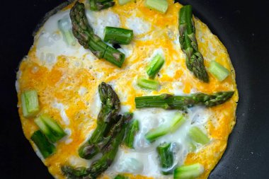 Parlak yeşil kuşkonmaz, mızraklar ve parçalar mükemmel bir şekilde altın, kabarık tavada omlet üzerine dizilmiştir. Bu mutfak sahnesi bahar esinlenilmiş bir kahvaltı ya da branç yemeğinin tazeliğini ve basitliğini vurgular, mevsimsel malzemeleri kutlar..