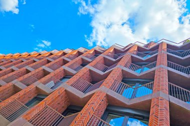 Canlı, mavi gökyüzüne karşı geometrik tuğla cepheli modern bir gökdelenin çarpıcı görüntüsü. Balkonların ve pencerelerin eşsiz deseni göz alıcı bir mimari tasarım, kentsel gelişim ve yenilik yaratır..