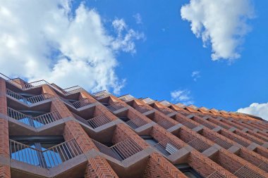 Modern tuğla bina, geometrik tasarım elementlerinin çarpıcı yükselişi. Mimari, çağdaş kentsel gelişim ve tasarımı vurgulayan tüylü beyaz bulutlarla noktalanmış canlı mavi gökyüzü ile çelişiyor..