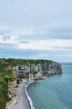 Kıyı kayalıkları kıyı şeridi boyunca belirgin bir şekilde yükselirken, yemyeşil alanlar kenarlarını kaplıyor. Huzurlu okyanus dalgaları sakin bir gökyüzünün altında, kumlu kumsalda nazikçe tur atar. Bu pitoresk deniz manzarası doğanın barışçıl güzelliğini temsil ediyor..