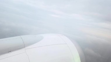 Bir uçak penceresinden, parlak beyaz bir kanadı, yumuşak ve dağınık bulutların arkaplanına karşı gösteren bir görüntü. Hava yolculuğunun ve keşiflerin huzur dolu sahnesi, uçuşun sükuneti ve enginliği..