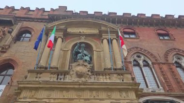Bologna, İtalya, 3 Ocak 2026: Avrupa bayraklarıyla çevrili bir oyukta heykel bulunan tarihi bir bina cephesi. Yapıda kemerli pencereler ve süslü taş işçiliği gibi ortaçağ mimari unsurlar sergilenirken, kültür mirasını ön plana çıkarıyor.