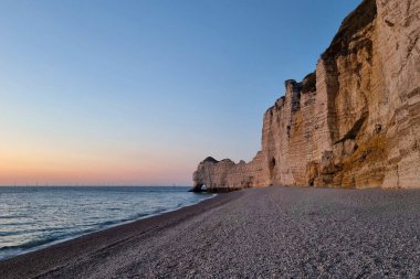 Cliffs rise along a pebbly beach with waves lapping at the shore. The sun sets on the horizon, casting a warm light over the scene. Wind turbines can be seen in the distance.