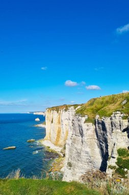 Yüksek kayalıklar okyanusun üzerinde kayalara çarpan dalgalarla dururlar. Parlak mavi bir gökyüzü uzaktaki kıyı manzarasını net bir şekilde gösteriyor..