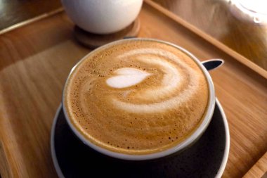 Karmaşık kalp şeklinde latte sanatı ile köpüklü bir latte, sıcak, davetkar bir kahve dükkanında ahşap bir tepside. Sütlü dokular ve lattenin zengin rengi bir rahatlama ve hoşgörü hissi uyandırıyor..