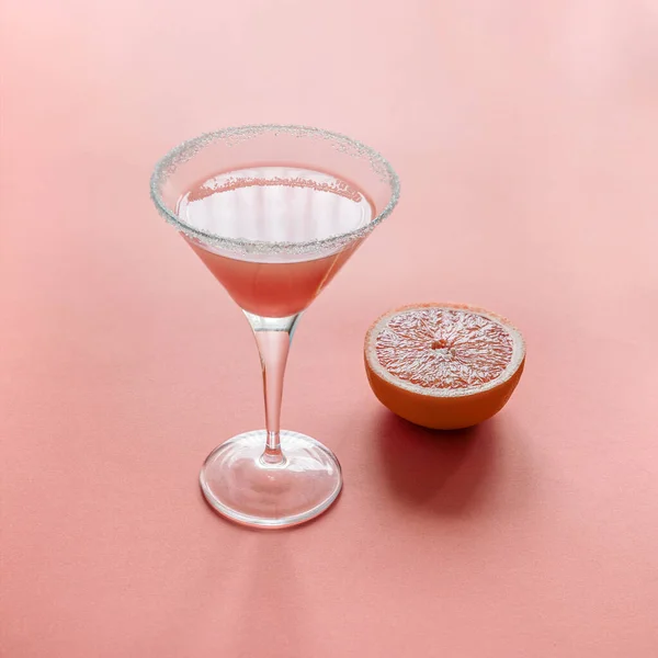 Cocktail glass with white sugar rim and refreshing grapefruit beverage. Reflection in the liquid, minimal summer concept on pink background.