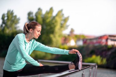 Genç ve aktif bir kadın sabah antrenmanı ve koşusu sırasında nehir kenarında bacak esnetiyor. Dinamik ve sağlıklı yaşam tarzı. 