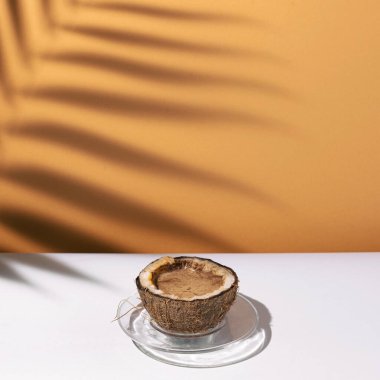 Doğal kakao ya da kremalı kahve hindistan cevizinin yarısına dökülüp küçük cam tabağa konmuş. Arka planda palmiye ağacı gölgesi var. Boşluğu kopyala