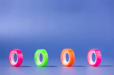 Küçük şeffaf selobant neon renklerde yuvarlanıyor. Kopyalama alanı olan mavi arkaplan. Okul ve ofis malzemeleri.