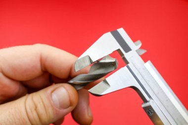 Bir kalibre çapını kırmızı arka planla karşılaştırırken, bir matkap ucu tutan el. Metalden yapılmış hassas bir enstrüman olan kalibre, net bir ölçü ölçeği içeriyor. Konferansta kesinlik ve ölçüm, DIY ve atölye faaliyetleri üzerinde duruluyor.