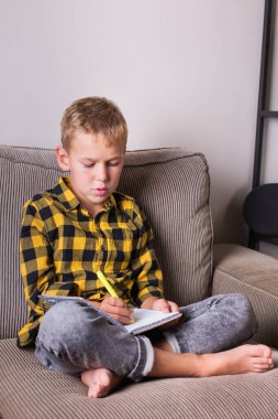 Kanepede oturan ve defterine yazan sevimli gülümseyen çocuk. Evde kal, kilitle, sosyal uzaklık, karantina.