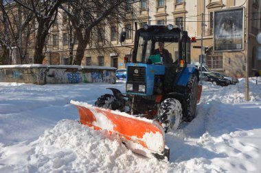 Kar temizleme traktörü döner fırçayla karları parktaki patikadan süpürüyor. Kepçe ve otomatik fırçalı kırmızı traktör karı kaldırır. Saban ile belediye karayolu temizleme aracı