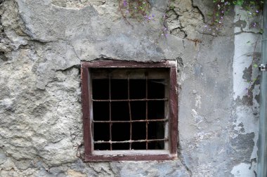 Terk edilmiş bir binanın duvarında metal parmaklıklar olan eski bir pencere. Çatlak alçı, pürüzlü doku ve karanlık açıklık kasvetli bir tarihi atmosfer yaratır..