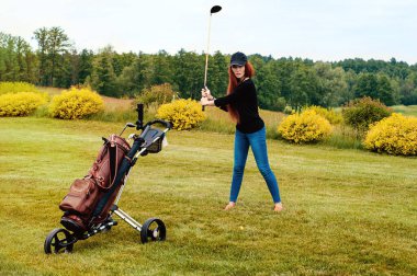 Golf sahasında güzel, kızıl saçlı bir kız oyuncu. Elinde golf sopalarıyla şık, kahverengi bir çanta var..