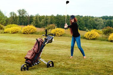 Golf sahasında güzel, kızıl saçlı bir kız oyuncu. Elinde golf sopalarıyla şık, kahverengi bir çanta var..