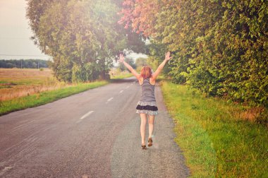 Kızıl saçlı kız yol boyunca yürüyor ve güneşin tadını çıkarıyor. Güneşli bir gün, ağaçlar, yeşil alan.