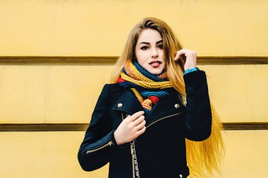 Sarı duvara karşı duran, kışlık elbiseli ve gülümseyen genç, güzel sarışın. Kış portresi.